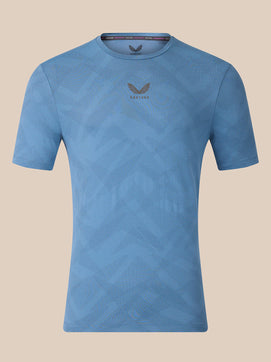 A light blue short-sleeve athletic t-shirt with a subtle geometric pattern and a dark gray wing logo with 'CASTORE' text on the center chest, displayed against a solid beige background.