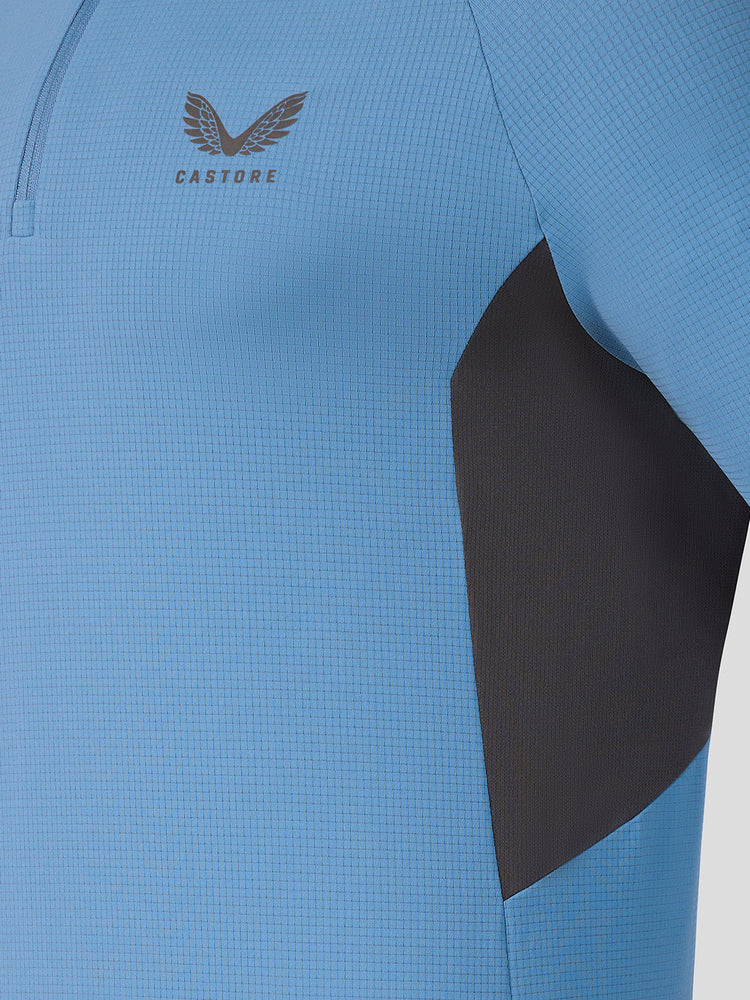 Close-up of a light blue athletic quarter-zip top with a dark gray side panel and a gray Castore logo on the chest.