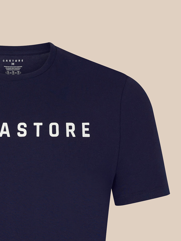 Close-up of a navy blue short-sleeve t-shirt with 'CASTORE' printed in white across the chest, shown against a solid beige background.