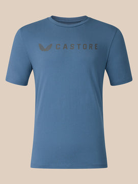 A blue short-sleeve crewneck t-shirt with a subtle dark gray Castore logo printed across the chest, displayed on a solid beige background.
