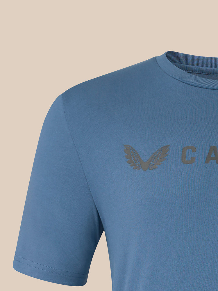 A close-up of a blue short-sleeve crewneck t-shirt featuring a tonal gray embroidered wing logo and the letters 'CASTORE' on the chest, set against a solid beige background.
