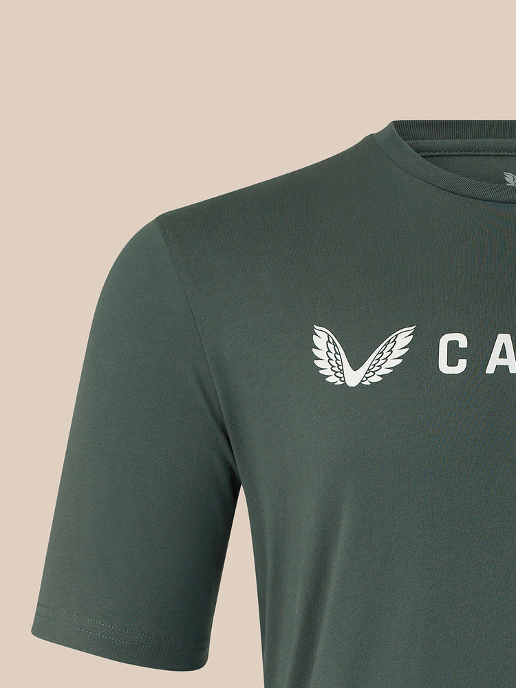 Close-up of a dark green short-sleeve t-shirt with a white winged logo and partial white text on the chest, set against a solid beige background.